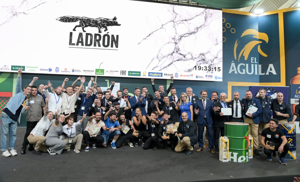 Large group of people posing together on a stage, smiling and raising hands, with a marble banner and sponsor logos in the background.