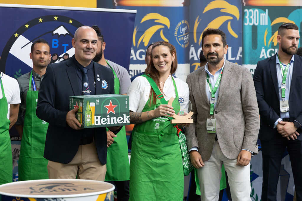 Group of award participants posing: man with Heineken case, woman with glass trophy, others in green aprons.