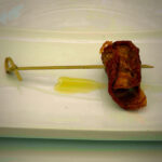 Wooden skewer rests on a white plate, with a crumpled brown dried leaf-like object wrapped around the right end and a pale yellow smear nearby.