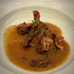 Braised lamb shank in a rich brown gravy, garnished with green herbs in a white bowl