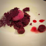 Pink ice cream scoop atop crumbled purple cake with a lacey wafer on a white plate and red sauce droplets nearby.