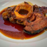 Braised beef short ribs in a rich brown-red sauce with a hollow marrow bone visible on a white plate