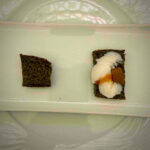Two small squares of dark bread on a light plate; the right piece is topped with white cream and orange jam/jelly, left piece plain.