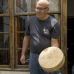 Man wearing glasses and a dark t-shirt stands outdoors, holding a large round wheel of cheese with a textured rind.