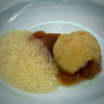 A fried breaded ball sits on a red sauce with bubbly, foamy liquid on a white plate.